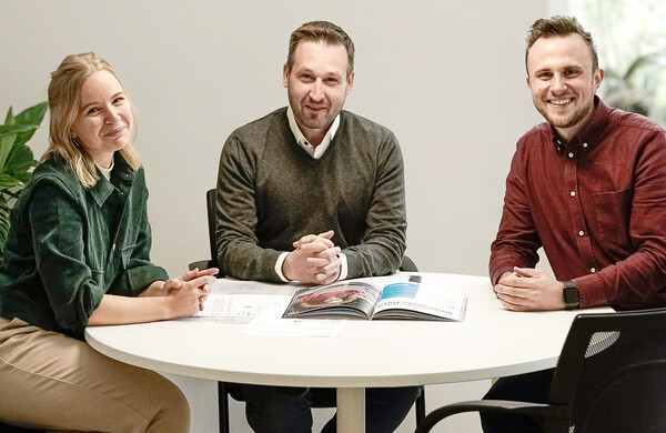 Jana Buhl, Christian Eßlinger und Dominik Stöcker sitzen in die Kamera schauend am Besprechungstisch.